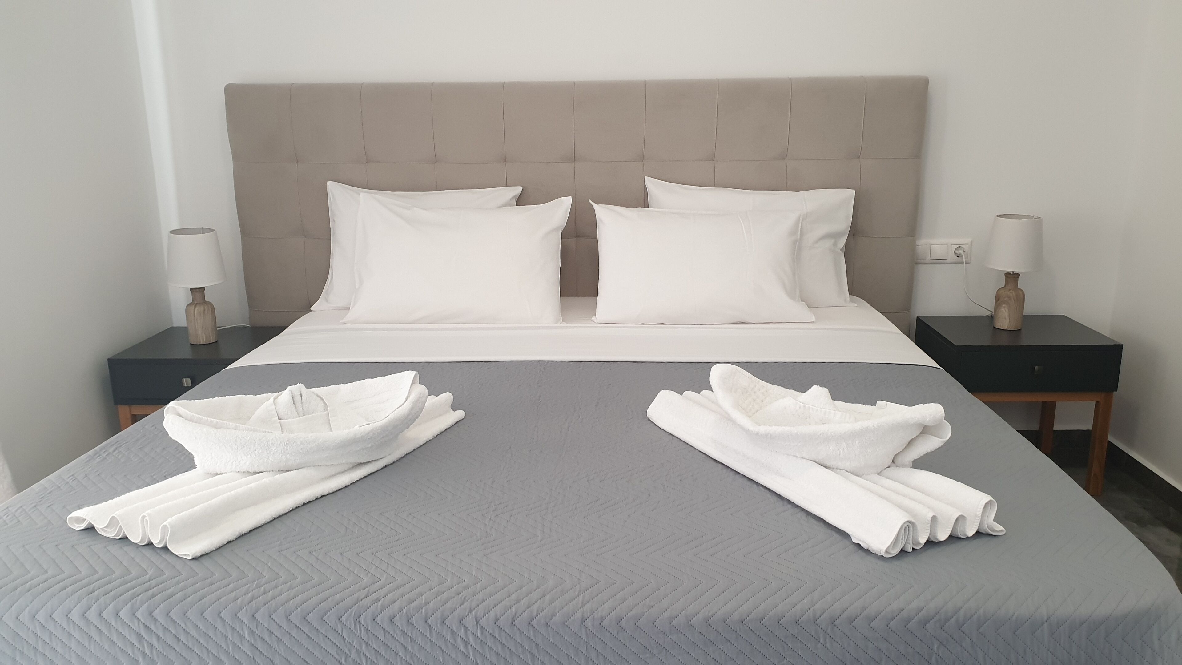 A neatly made bed with a grey quilt, two white pillows, and two rolled white towels on the bed.