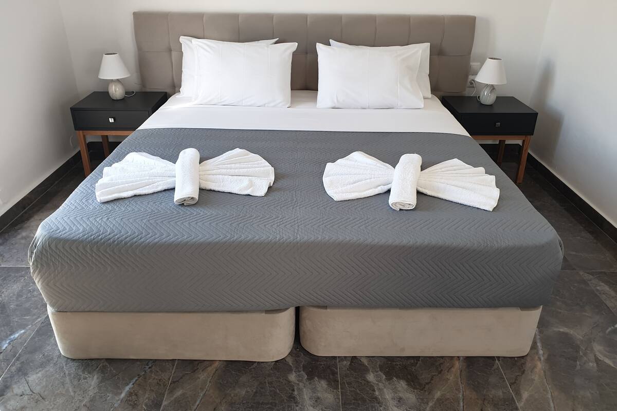 Cama bem arrumada com duas toalhas brancas dobradas sobre o cobre-leito, duas mesas de cabeceira com abajures e uma cabeceira grande e acolchoada.