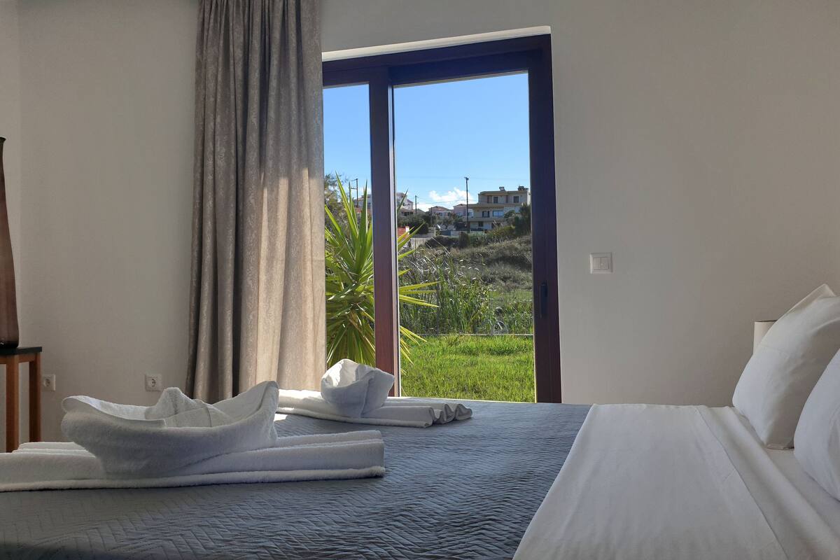 Quarto de hotel com cama, janela com vista para o exterior, um vaso em uma mesinha de cabeceira e toalhas sobre a cama.