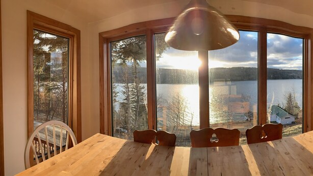 Un comedor con una mesa grande y sillas, ventanas amplias con vista a un lago y un bosque, y una lámpara colgante.