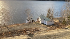 A lakeside cabin with a small deck and a covered outdoor area, surrounded by trees and a dirt path. - Rangeley Waterfront Home Retreat (Rangeley Plantation)