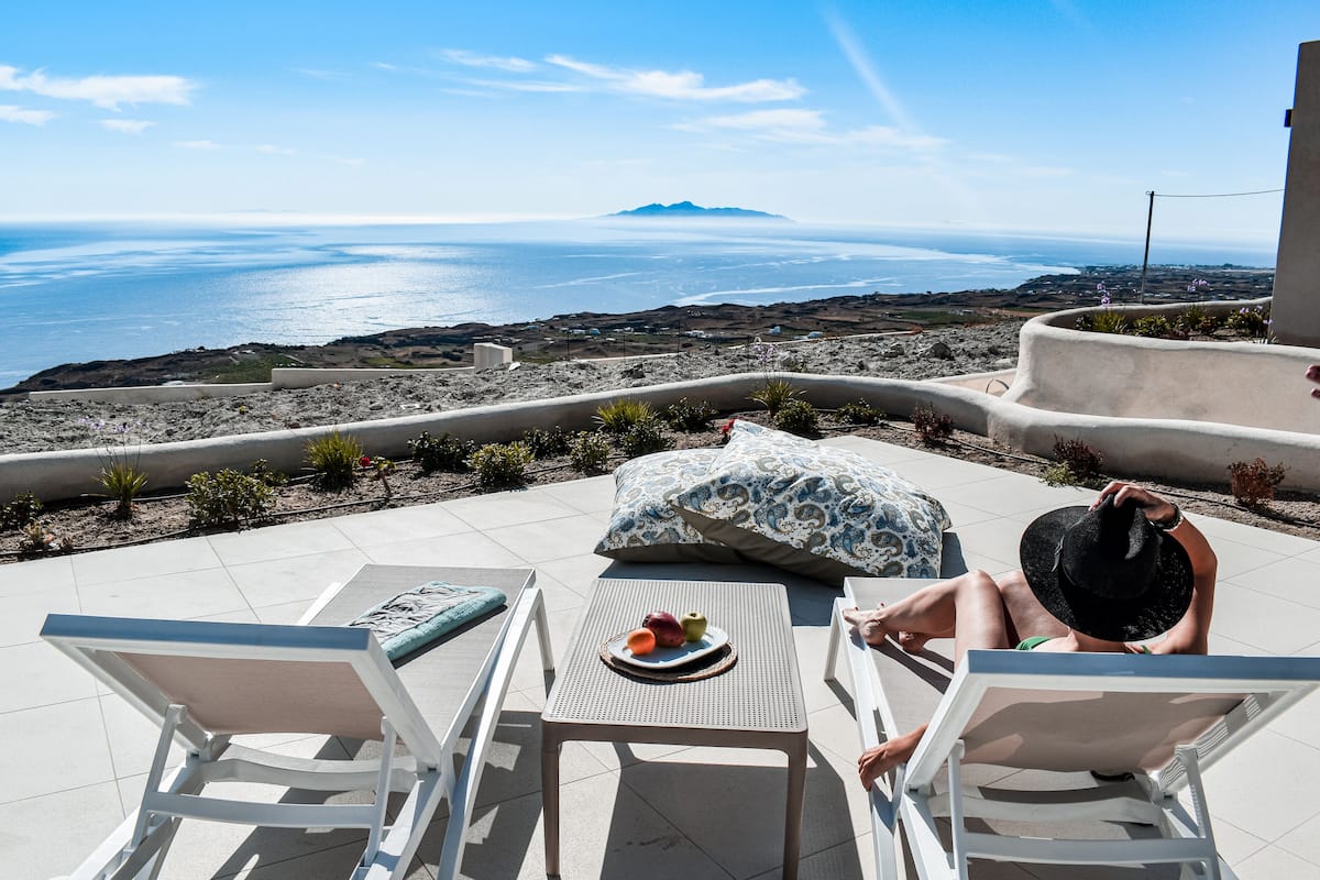 A terrace with two lounge chairs, a table with fruits, and a view of the ocean and distant mountains.