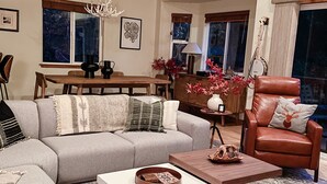 A spacious living room with a large sectional sofa, a wooden coffee table, and a leather armchair. A dining table with chairs is visible in the background, and a guitar is mounted on the wall.