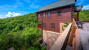 A log cabin with a stone foundation, a balcony, and a view of a forested hillside. - Legacy Mtn Resort Cabin w/ Mtn View - 2 King Beds + 2 Sofa Beds HotTub & Jacuzzi (Sevierville)