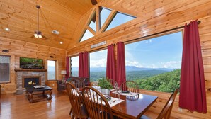 A wooden cabin interior with a dining table, chairs, a fireplace, and large windows offering a mountain view. - Legacy Mtn Resort Cabin w/ Mtn View - 2 King Beds + 2 Sofa Beds HotTub & Jacuzzi (Sevierville)