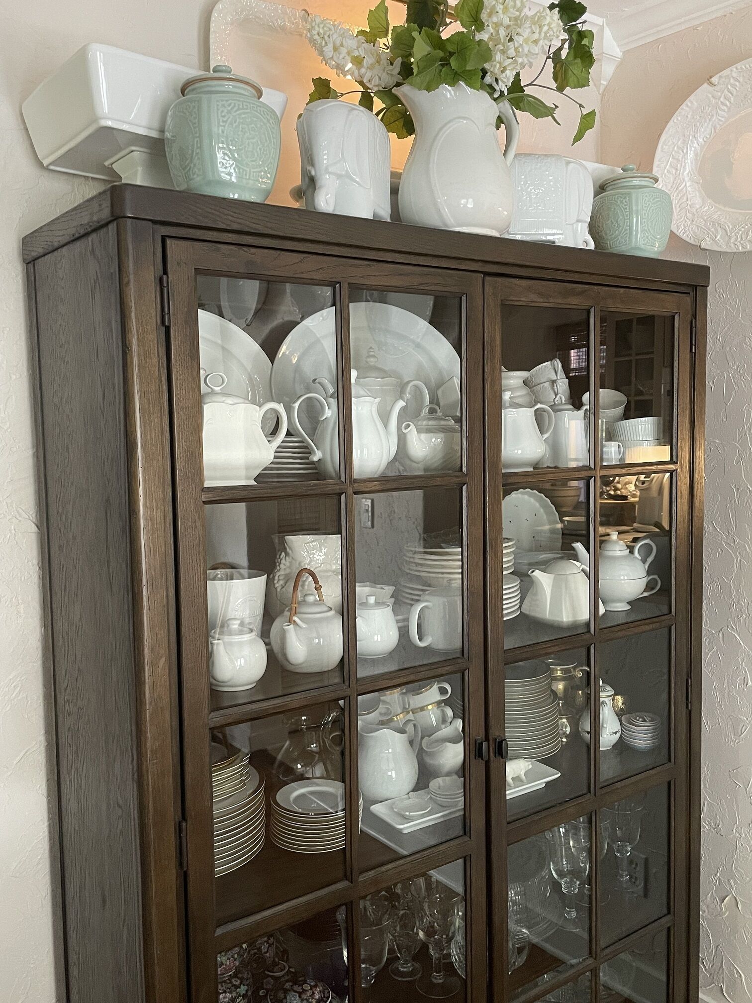 Ein Holzschrank mit Glastüren, der weiße Keramikteller, Teekannen und Gläser beherbergt.