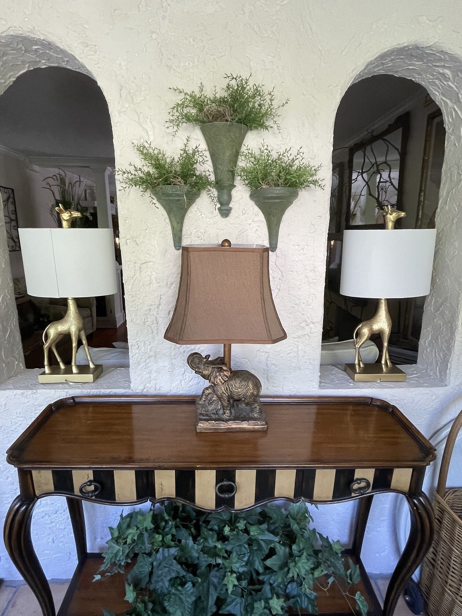 A wooden table with a lamp and a decorative elephant figurine. There are two wall-mounted lamps with animal figurines and green plants above them.