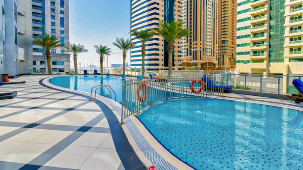 A swimming pool area with a tiled deck, palm trees, and high-rise buildings in the background. - HiGuests - The Marina Torch (Dubai)