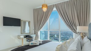 A hotel room with a large bed, a flat-screen TV, a desk with a chair, and a view of a cityscape through large windows. - HiGuests - The Marina Torch (Dubai)
