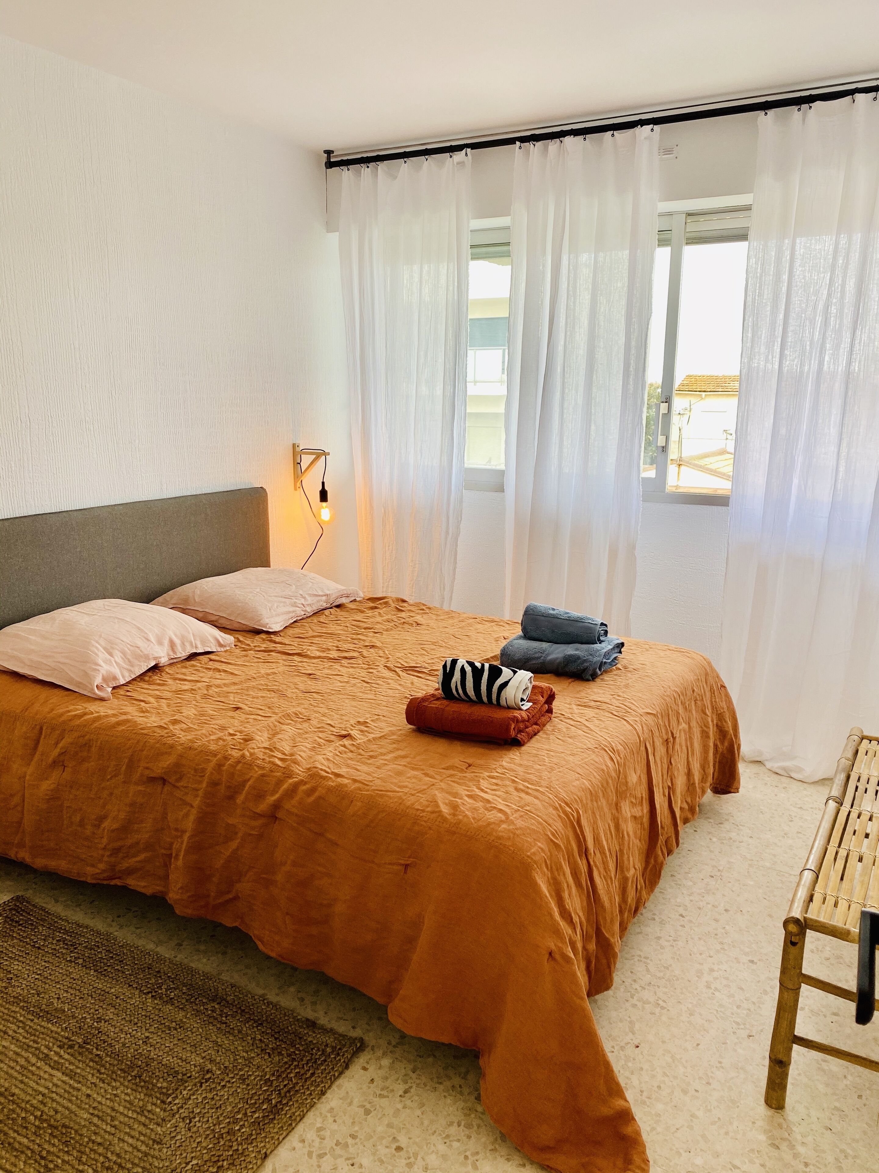 Quarto com uma cama grande, mesa de cabeceira, abajur e vista para o exterior através de cortinas translúcidas.
