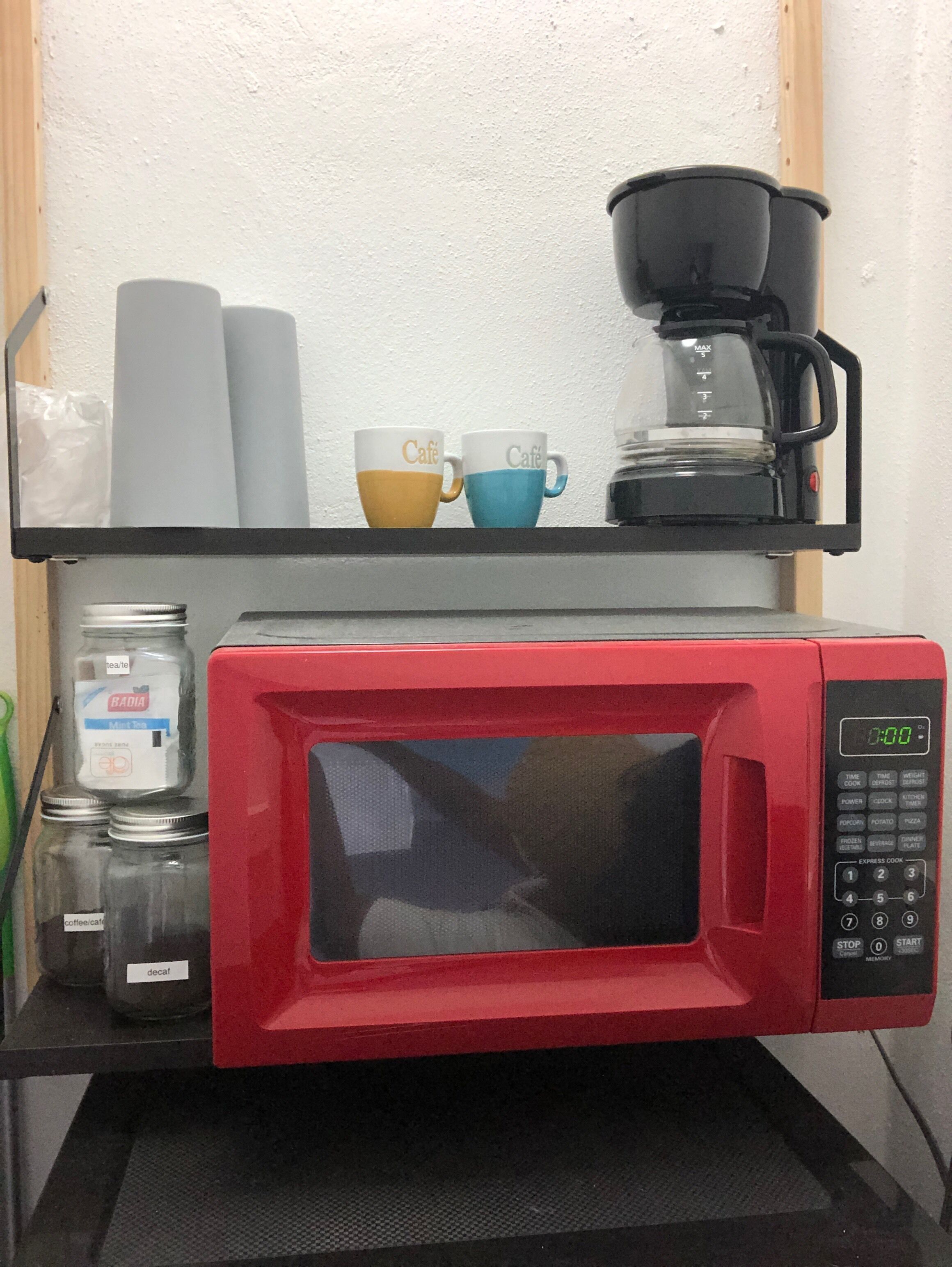 A red microwave on a shelf with two coffee cups and a coffee pot.