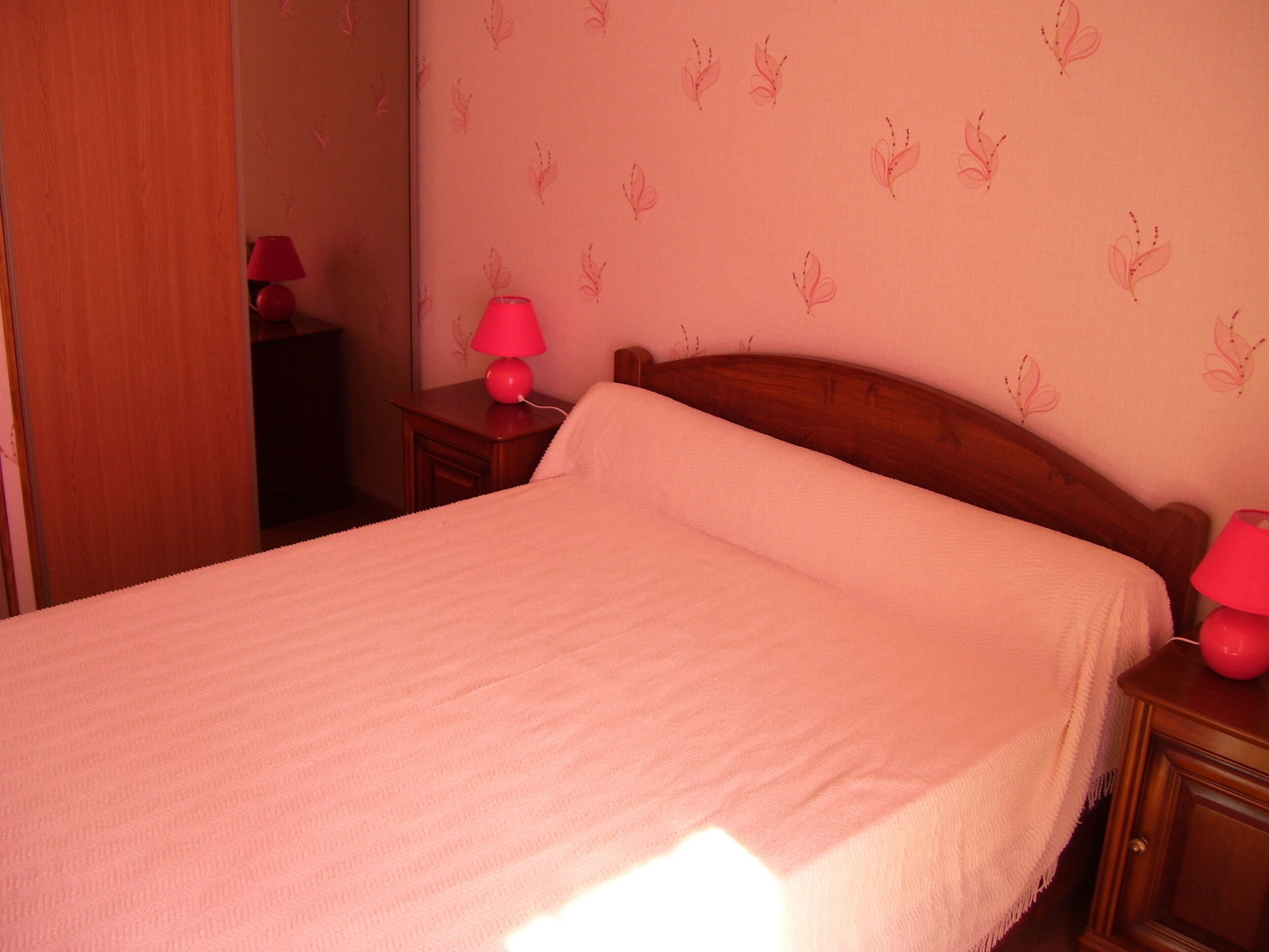 Un lit bien fait, recouvert d’une couvre-lit blanche et agrémenté d’oreillers, une tête de lit en bois, une table de chevet avec une lampe rouge et un mur orné d’un motif discret représentant des feuilles.