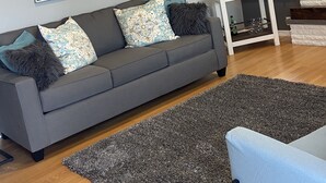 A modern living room with a grey sofa, patterned cushions, a small white side table, and a large painting on the wall.