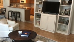 A modern living room with a sofa, coffee table, and TV stand. There is a kitchen area with wooden cabinets and a microwave.