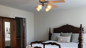 A bedroom with a wooden four-poster bed, a wooden chair with cushions, a ceiling fan, and a large mirror on a wooden cabinet.