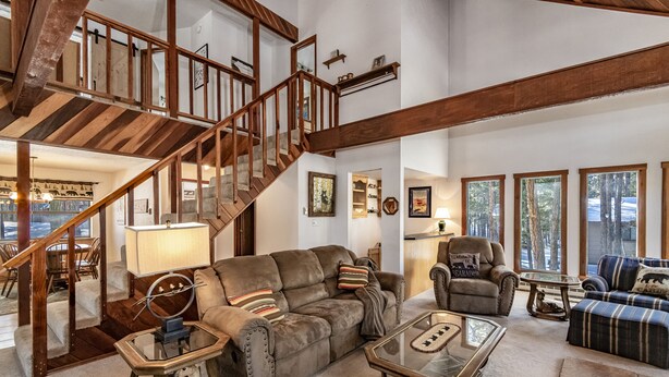 A spacious living room with a wooden staircase, a leather sofa, a glass coffee table, and a view of the outdoors through large windows. - Spacious Angel Fire Cabin Near Skiing, Hiking, and Fishing (Angel Fire)