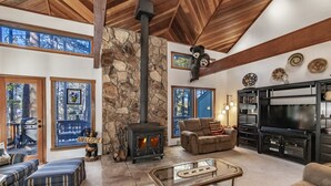 A cozy living room with a stone fireplace, comfortable seating, a television, and a large window overlooking a deck. - Spacious Angel Fire Cabin Near Skiing, Hiking, and Fishing (Angel Fire)