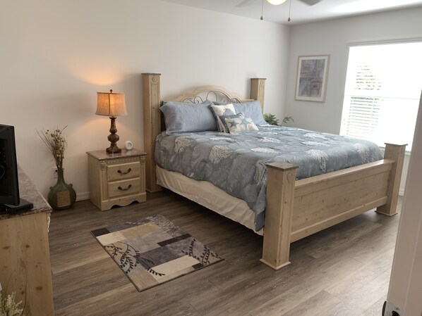 Un dormitorio con una cama de madera, una mesita de noche con una lámpara, una pequeña mesa con un jarrón y una ventana con persianas.