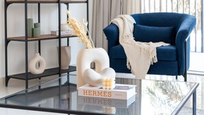 A living room with a glass coffee table, a blue armchair, a black bookshelf, and a window with curtains. - HiGuests - Boulevard Crescent Tower 2 (Dubai)