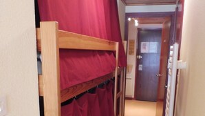 Un couloir étroit avec des lits superposés et des rideaux rouges, un plancher en bois et une porte au fond.