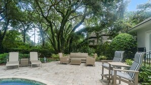 An outdoor patio with wicker furniture, a pool, and lush greenery.