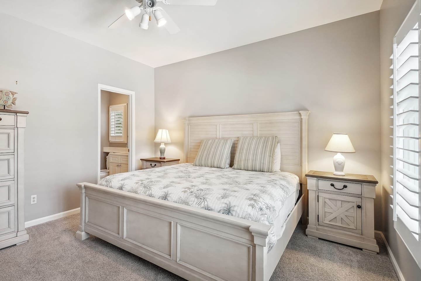 Une chambre à coucher avec un lit, des tables de chevet, un ventilateur de plafond et une fenêtre avec des stores.