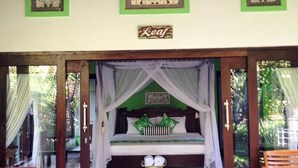 A room with a bed, a small table, and a chair. There are curtains on the windows and a sign on the wall. - Kirana Homestay - Leaf Bungalow (Karangasem)