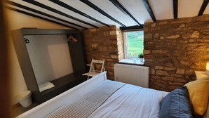 A bedroom with a stone wall, a window, a bed with pillows, a hanging chair, and a small table with a lamp. - Cottage with Large Drive (Wiltshire)