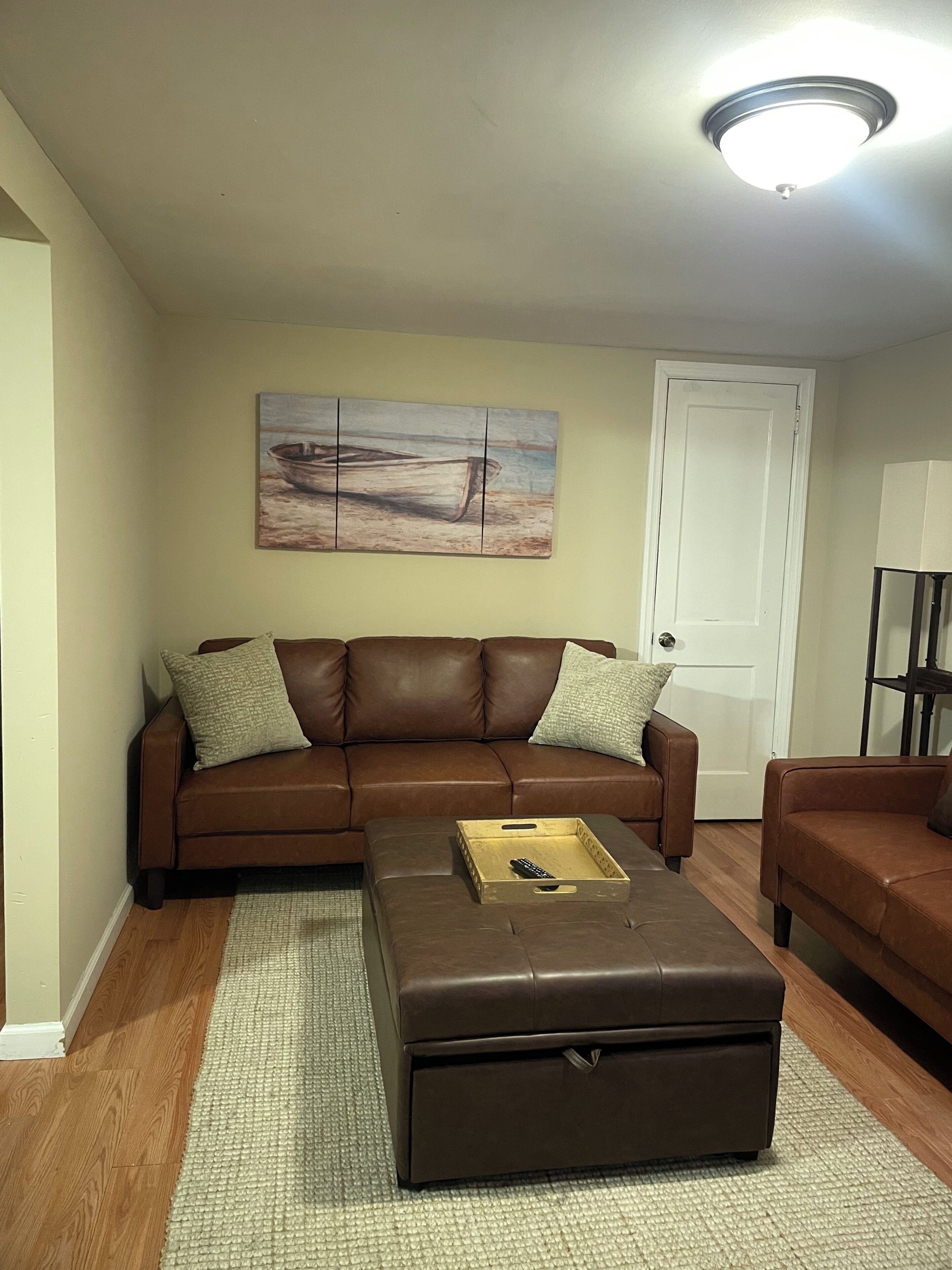 A living room with a brown leather sofa, a wooden coffee table, a painting on the wall, and a ceiling light.