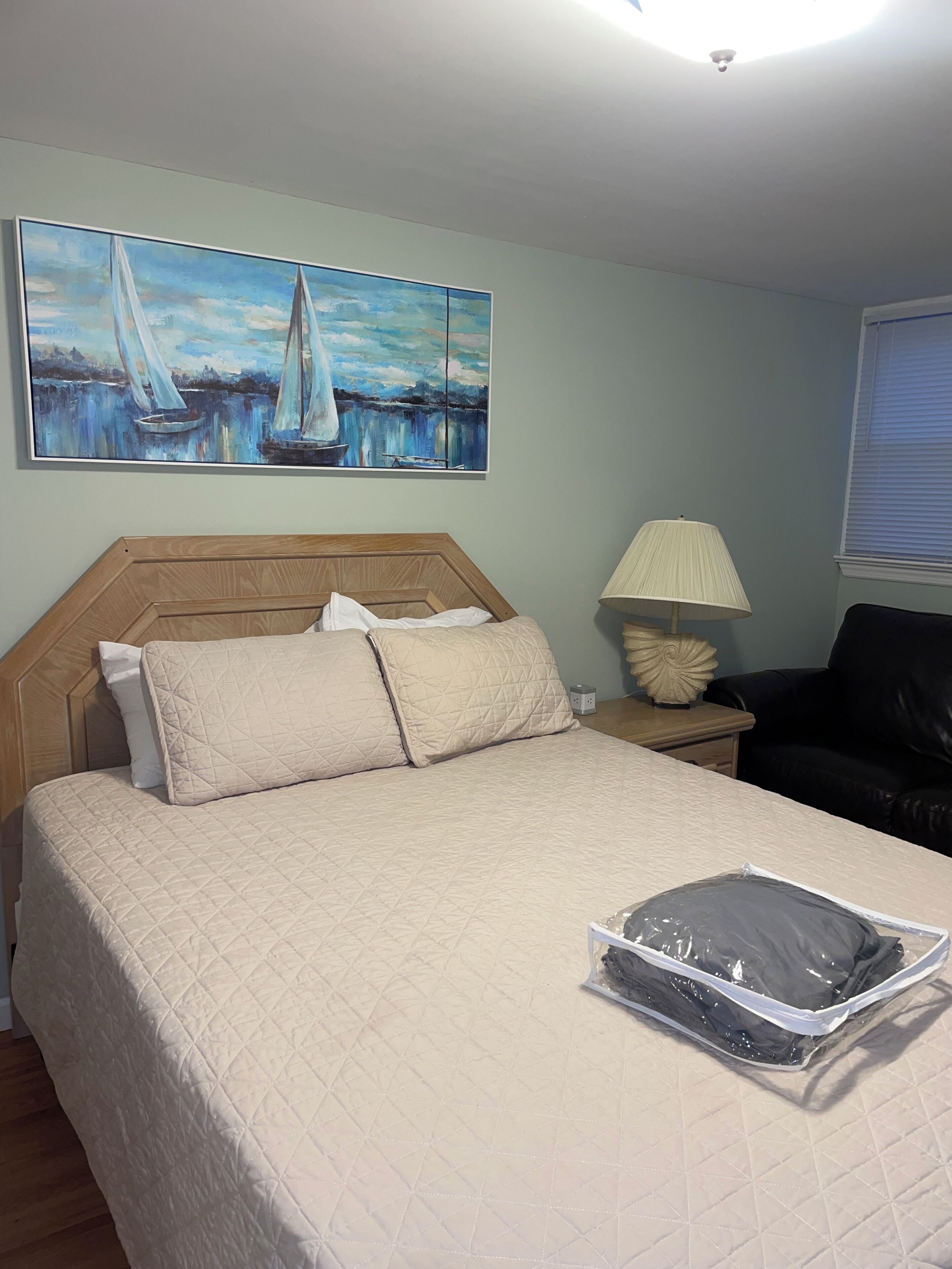 A hotel room with a bed, a painting of sailboats, a lamp, and a chair.