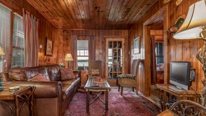 L’intérieur chaleureux d’un chalet avec un canapé en cuir, un fauteuil en osier, une table basse en bois et un téléviseur posé sur un meuble en bois.
