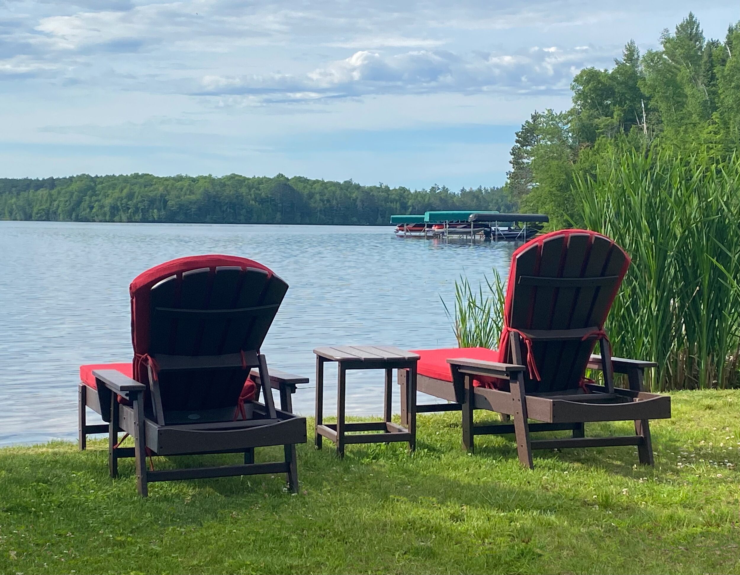 Two Adirondack chairs facing a lake with a dock and boats in the background.