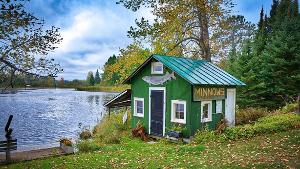 Une cabane verte avec un toit en métal, située au bord d’un lac, entourée d’arbres et agrémentée d’un banc.