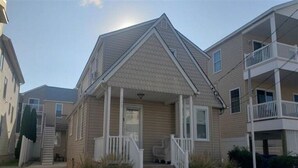 A two-story residential building with a gabled roof, a front porch with white railings, and a small front yard with landscaping.