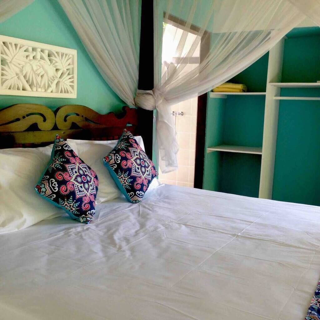 Een bed met witte lakens en twee kleurrijke kussens, een houten hoofdbord, een wit kunstwerk aan de muur, en een groene muur.