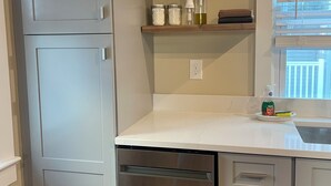 Une cuisine avec des meubles gris, un lave-vaisselle, un plan de travail sur lequel se trouve une bouteille de liquide vaisselle, et une fenêtre avec des stores.