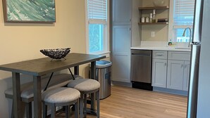 Une cuisine moderne avec une table à manger en bois, deux tabourets de bar, un réfrigérateur en acier inoxydable et une fenêtre avec des stores.