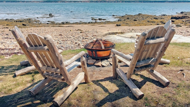 Two Adirondack chairs facing a body of water with a fire pit between them.