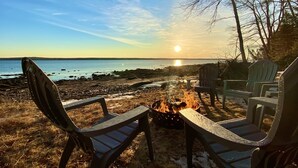 A beach scene with a fire pit and chairs facing the ocean.