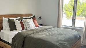 A bedroom with a wooden bed, white walls, a nightstand, and a window with a view of trees.