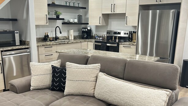 A modern living room with a sectional sofa, a dining area, and a kitchen with stainless steel appliances.