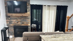 A modern living room with a sectional sofa, a flat-screen TV mounted on a wooden wall, a fireplace, and a kitchen area with a granite countertop.