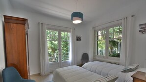 Une chambre avec deux lits, une armoire en bois, une chaise bleue et de grandes fenêtres avec des rideaux blancs.
