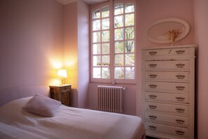 A bedroom with a bed, a dresser with multiple drawers, a radiator, and a window with a view of trees.