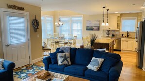 Una sala de estar con sofá azul, un comedor con mesa y sillas, y una cocina con refrigerador y electrodomésticos en la encimera.