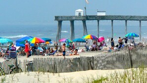 Escena de playa con un muelle de madera, sombrillas y gente relajándose en la arena.