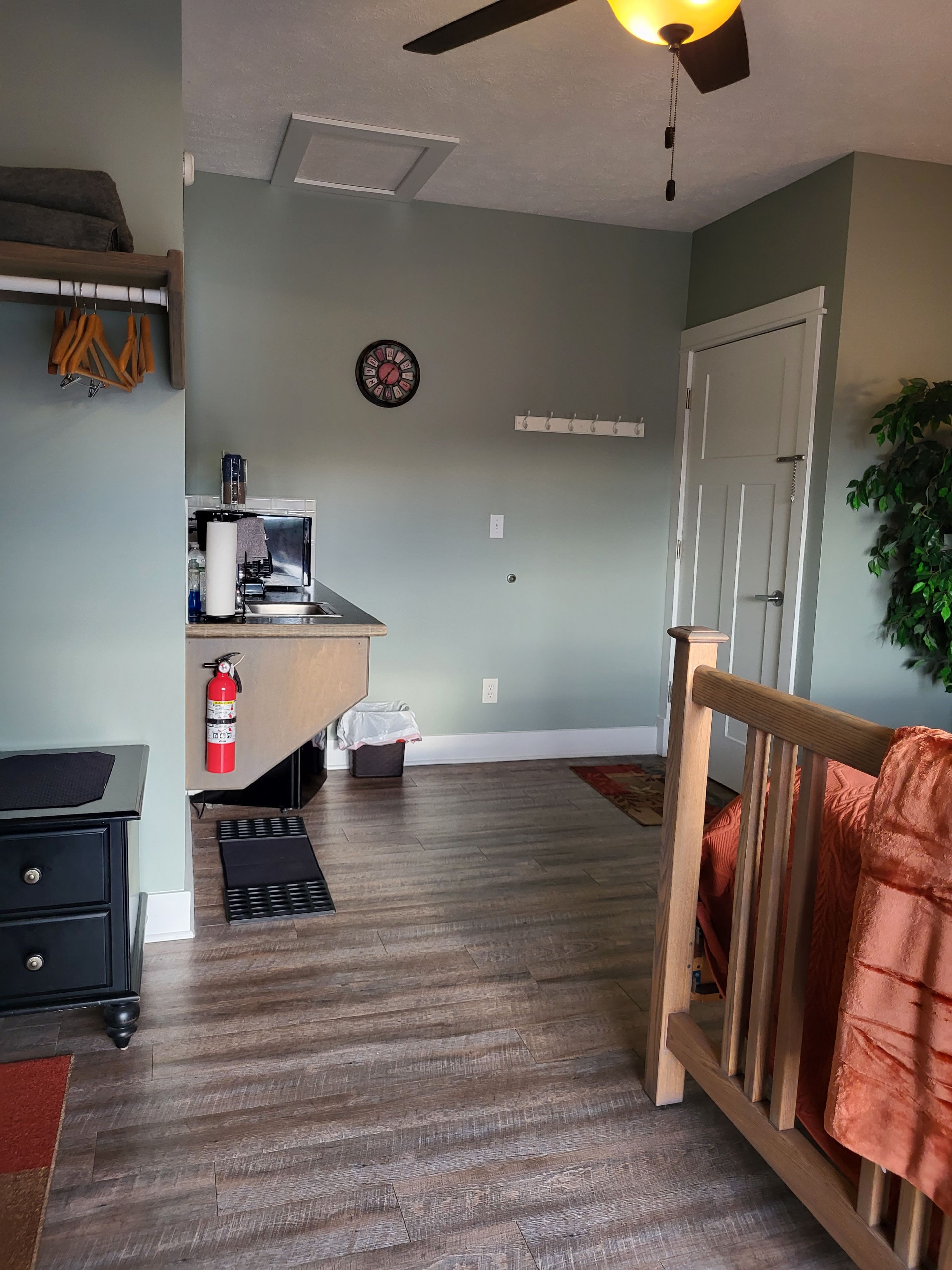 A room with a kitchenette, a ceiling fan, a clock, and a coat rack.