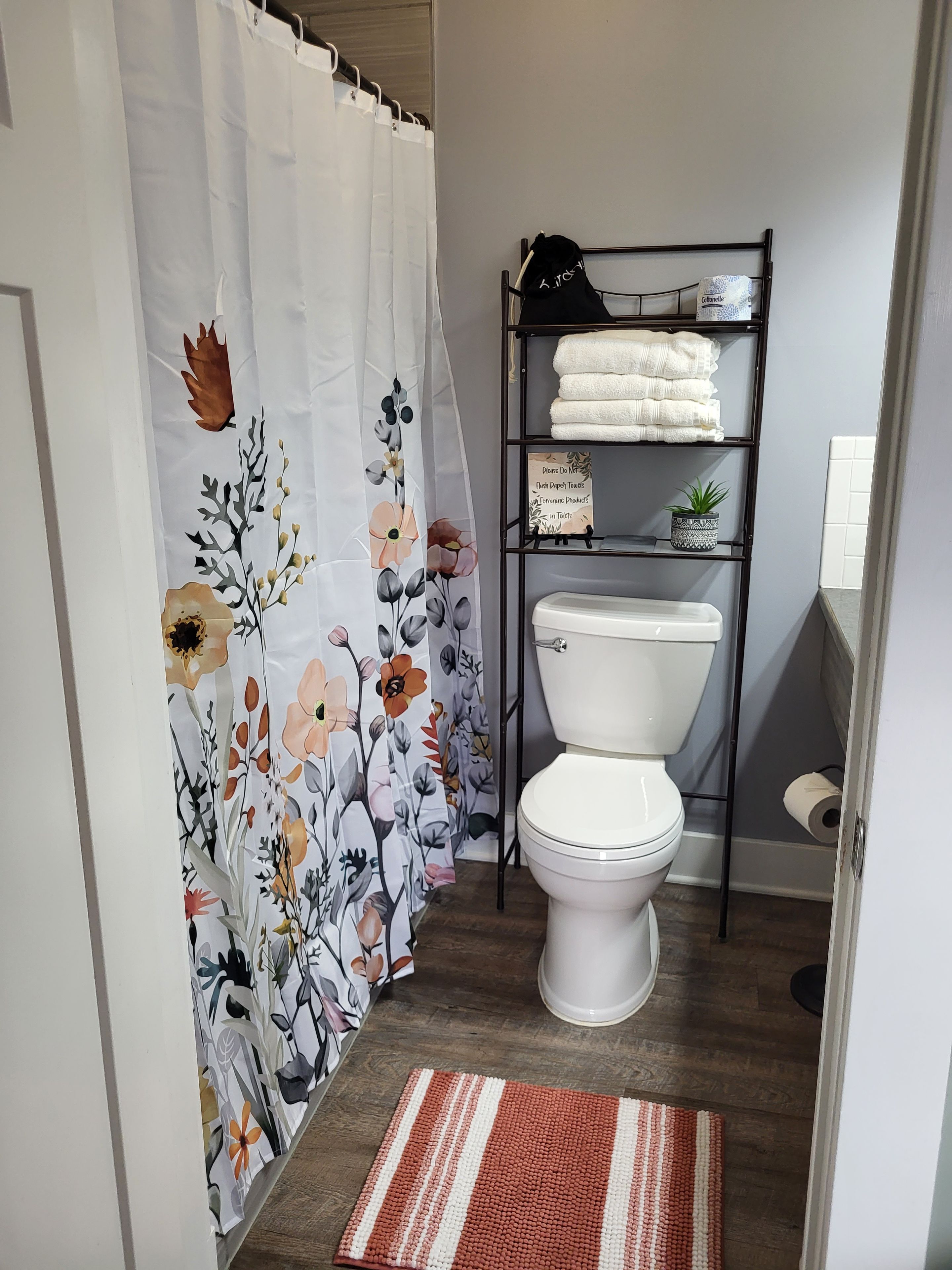 A bathroom with a toilet, a shelf with towels, a shower curtain with floral patterns, and a wooden floor.
