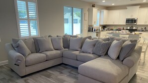 A modern living room with a L-shaped sectional sofa, a kitchen with white cabinets, and a large window providing natural light.