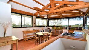 Una cocina con vigas de madera, un comedor con una mesa redonda y sillas, y ventanas amplias que ofrecen vistas al exterior.